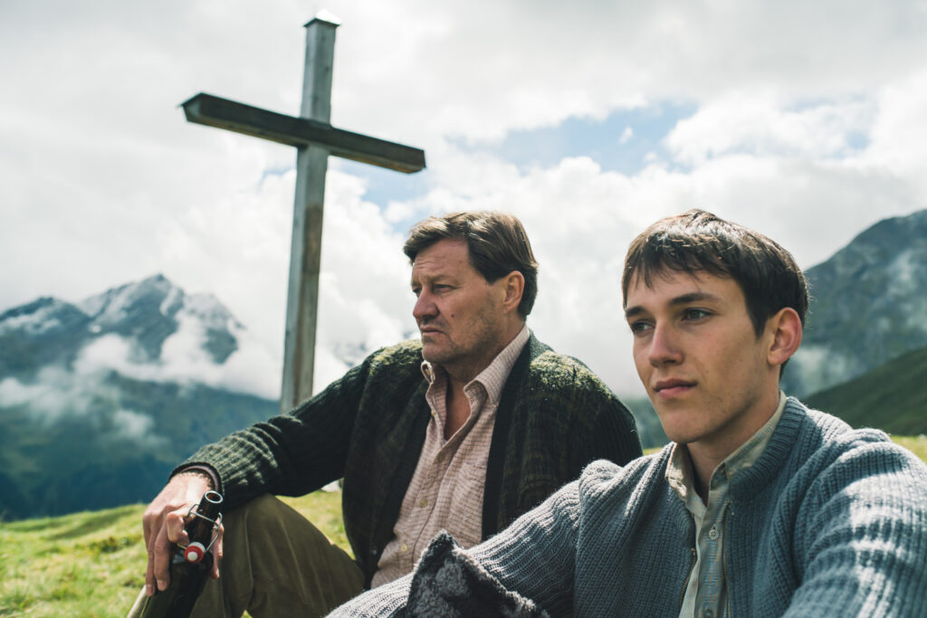 Vater (Harald Windisch) und Sohn (Jakob Mader) sitzen vor dem Gipfelkreuz in der Sonne und schauen in die Ferne. Der Vater hat eine Bierflasche in der Hand. 
Im Hintergrund das Gipfelkreuz und dahinter von Wolken umgebene Berggipfel .