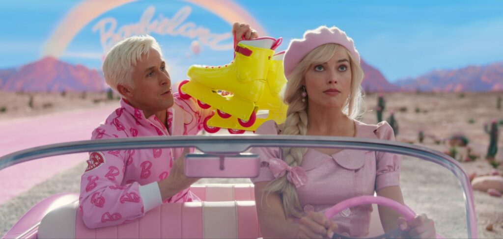 Ken und Barbie in einem Cabrio. Ken sitzt auf der Rückbank und hält ein Paar gelbe Rollerskates hoch. Im Hintergrund ist ein Regenbogen und der Schriftzug "Barbieland" zu sehen.
