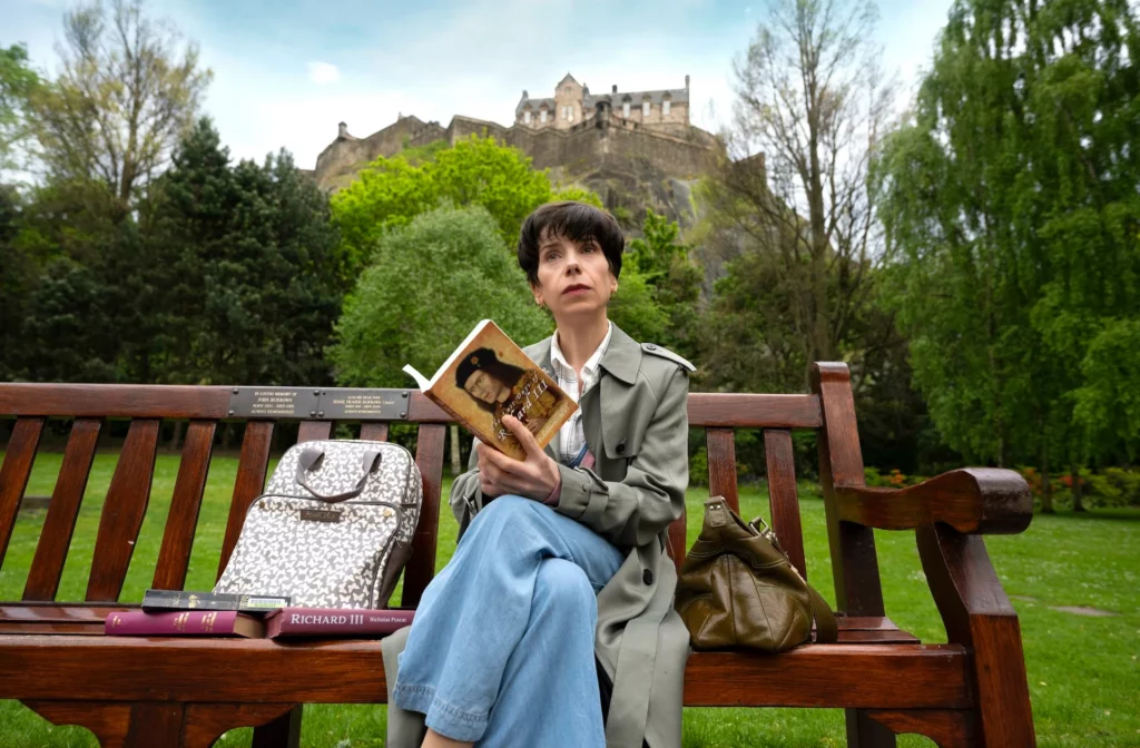 Sally Hawkins sitzt auf einer Parkbank mit einem Buch über Richard III in der Hand. Im Hintergrund befindet sich eine Burgruine. 
Filmstill aus THE LOST KING von Stephen Frears.